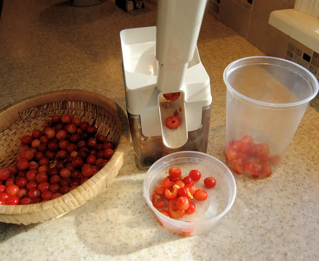 a basket of sour cherries, a cherry pitter and two plastic containers on a kitchen counter.