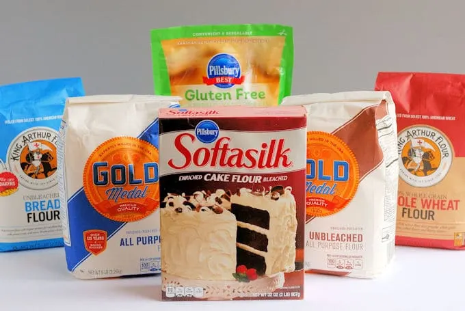 Six different bags of flour arranged on a white surface against a gray background.