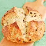 A loaf of Irish soda bread with raisins is set on a wooden cutting board with a green background.