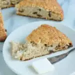 One wedge of date and spice scone on a white plate with a pat of butter and butter knife. another scone in the background