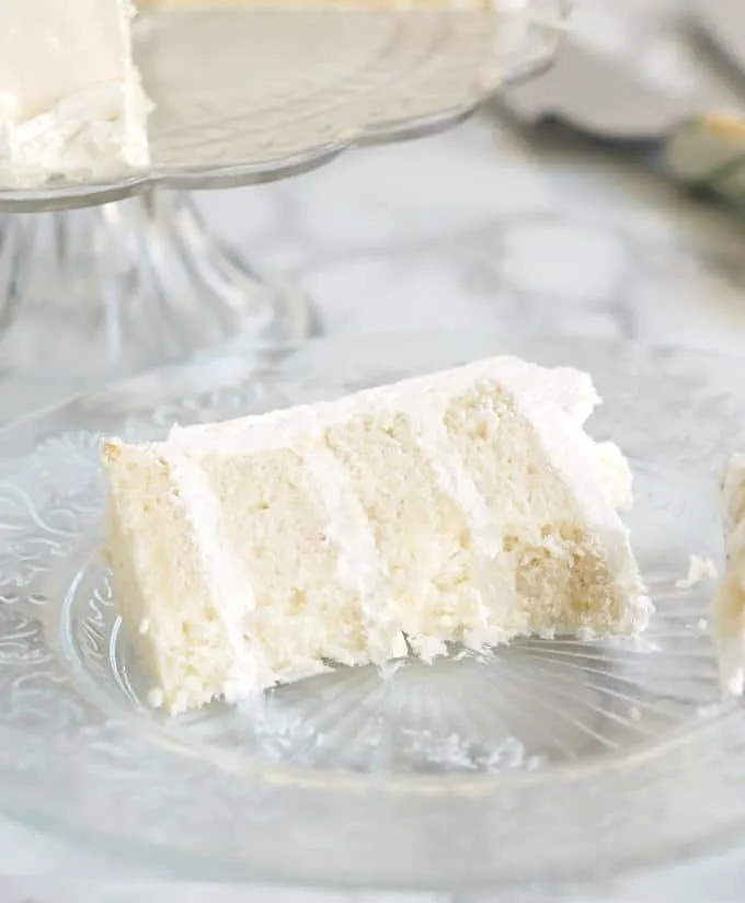 A half-eaten slice of four layer velvety soft white cake on a glass plate
