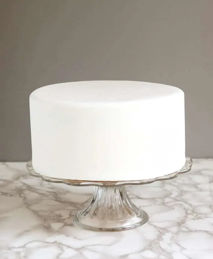 A white cake covered in rolled fondant on a glass cake plate