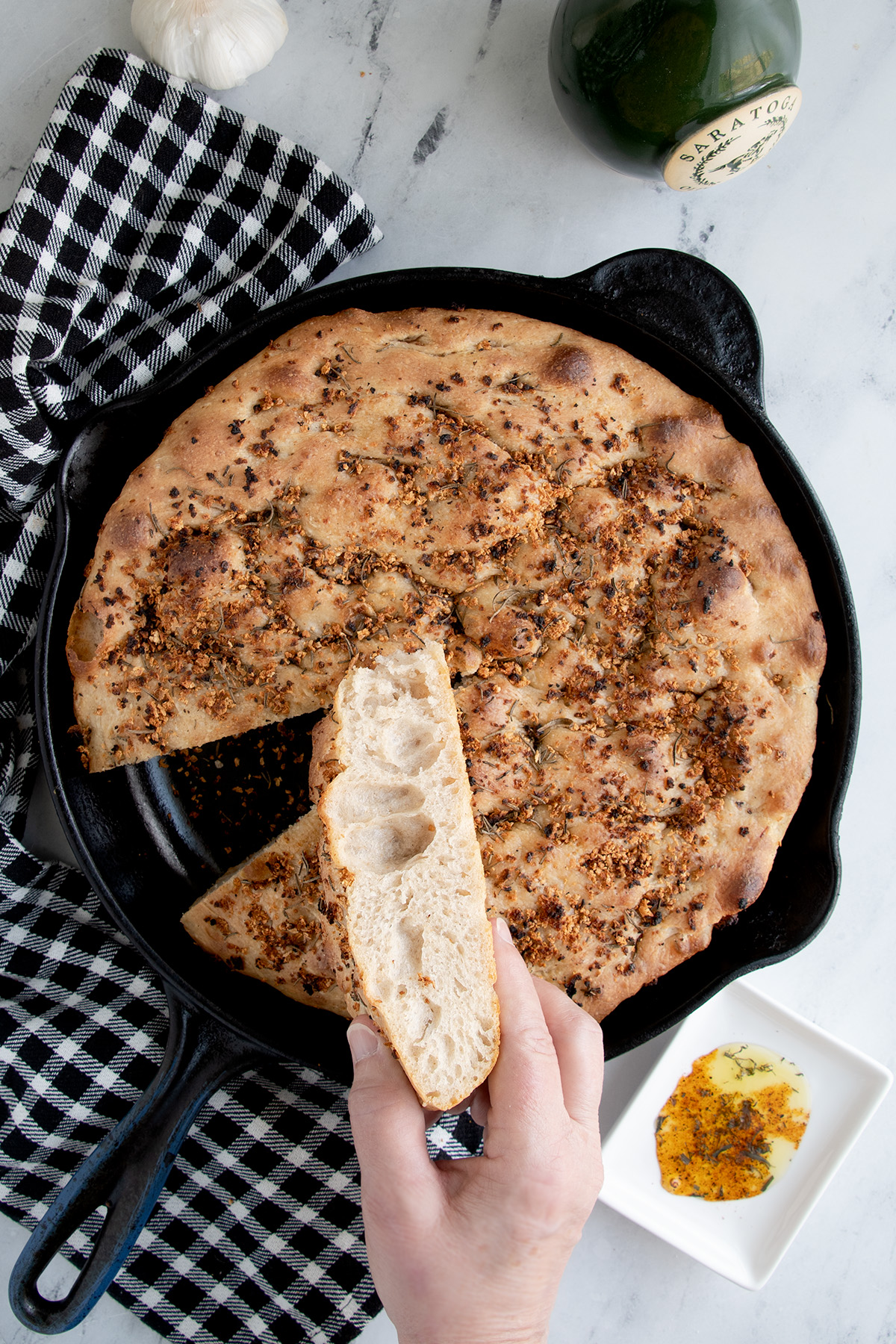 a focaccia in a skillet with a slice held by a hand.
