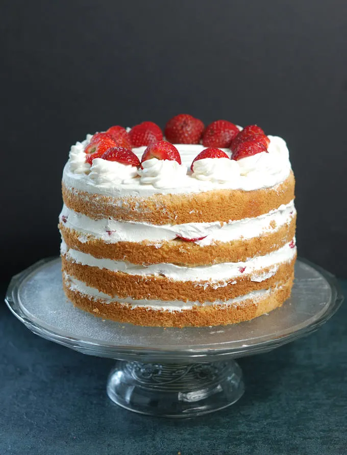 a four layer strawberry tall cake on a glass cake stand with a black background