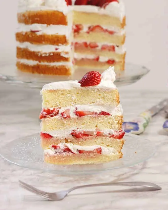 a slice of strawberry tall cake on a glass plate. A whole Strawberry chiffon cake in the background