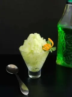 a bowl of green melon granita with an orange twist against a black background