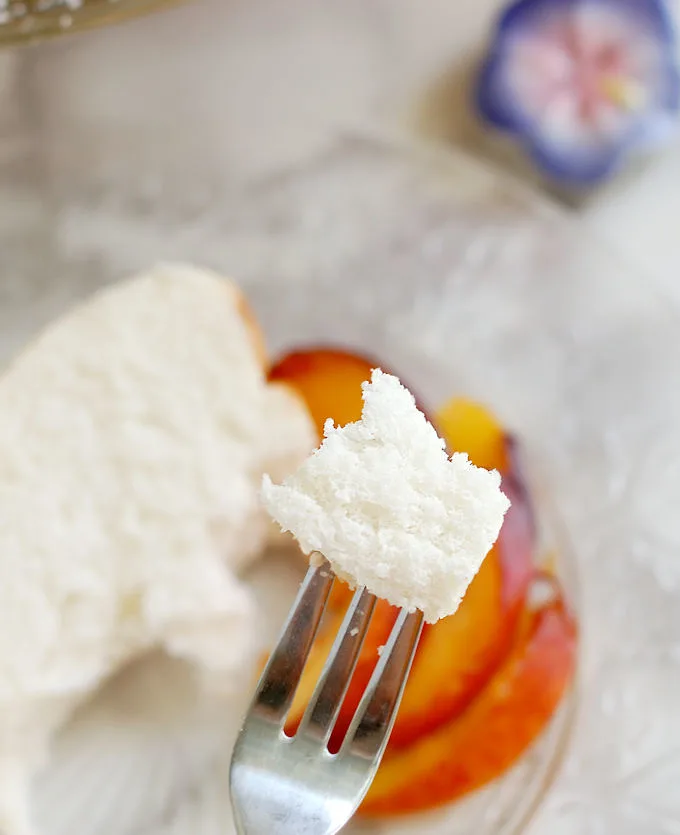 a closeup of a bite of angel food cake on the end of a fork with fresh peaches blurred in the background