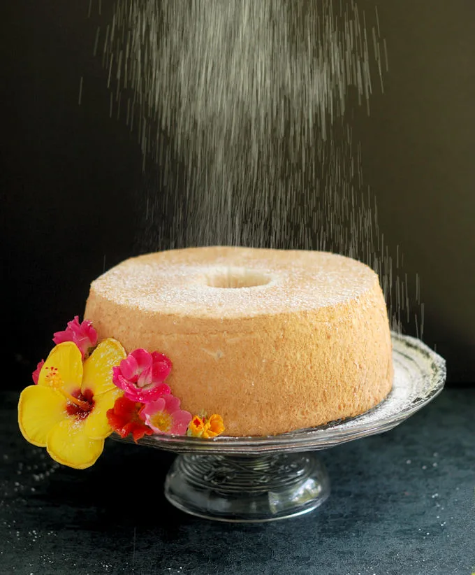 an angel food cake on a glass cake stand is being showered with powdered sugar.