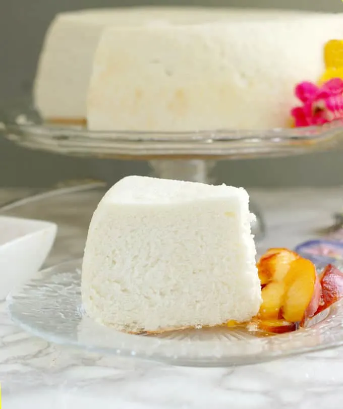 a slice of angel food cake on a glass plate with fresh peach slices. A whole cake is on a stand in the background