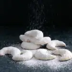 a pile of vanilla kipferl cookies on a black background with sugar pouring down
