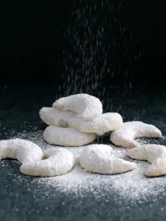 a pile of vanilla kipferl cookies on a black background with sugar pouring down