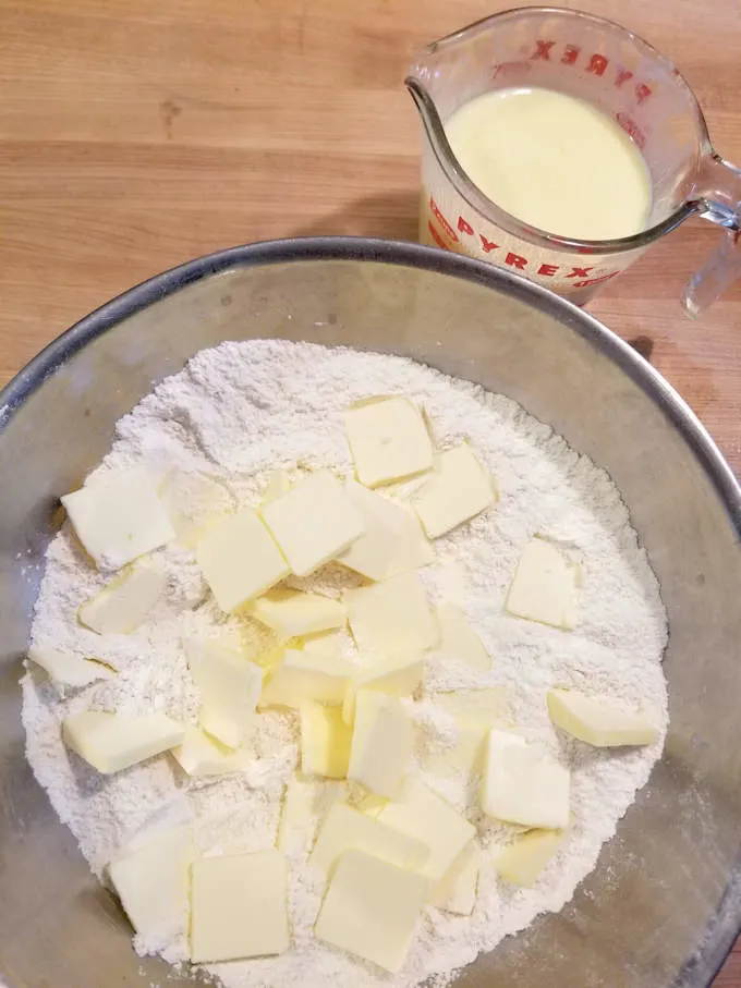 a bowl of flour with chunks of butter on top with a cup of buttermilk on the side.