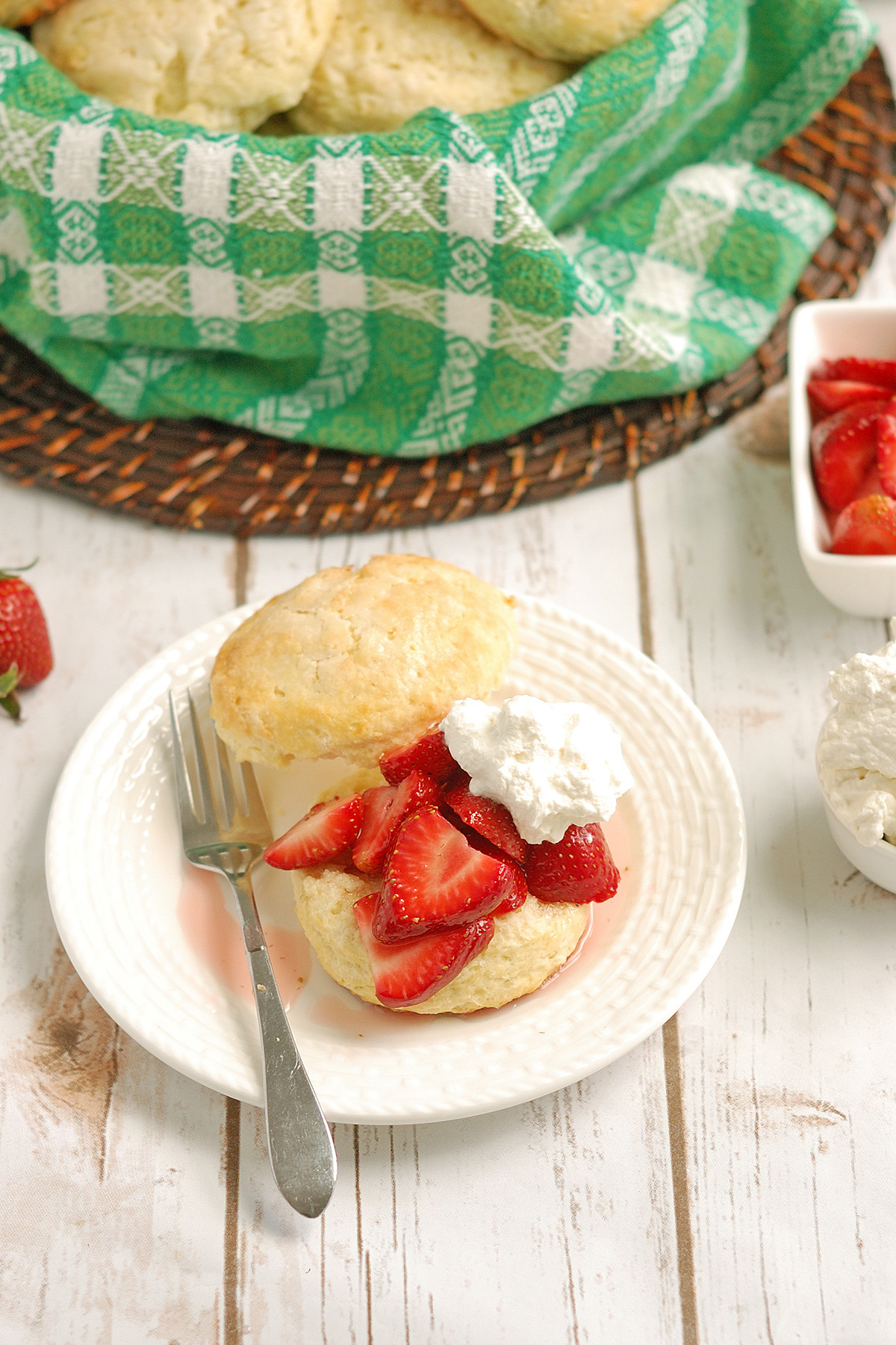 a strawberry shortcake on a plate. A basket of shortcake biscuits with a green cloth.