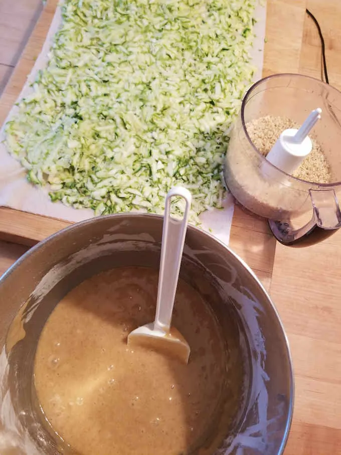 A bowl of cake batter, a tray of shredded zucchini and ground walnuts are the components for zucchini cake.