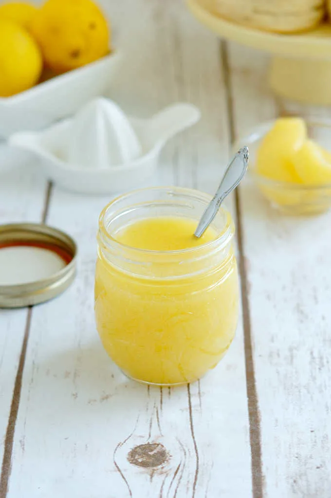 a jar of lemon curd on a table surrounded by lemons and scones.