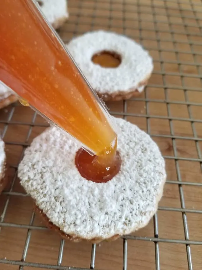 a piping bag filling a pistachio linzer cookie with apricot preserves