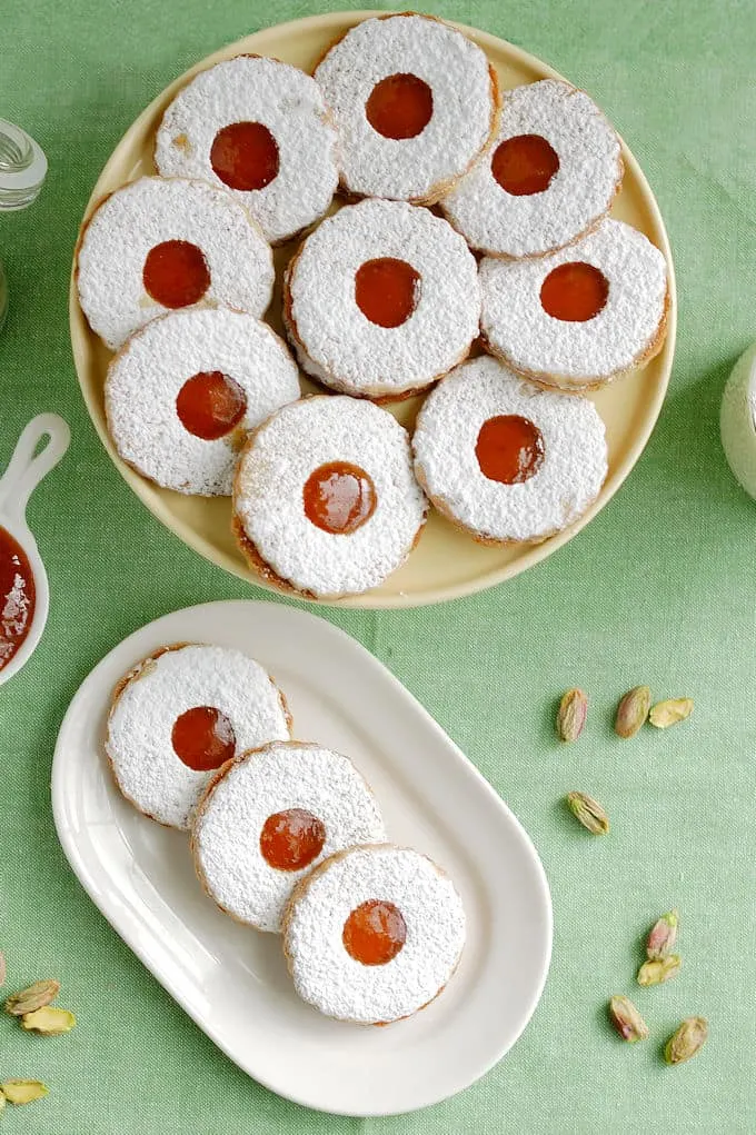 a plate of pistachio apricot linzer cookies in front of a cake stand with more cookies