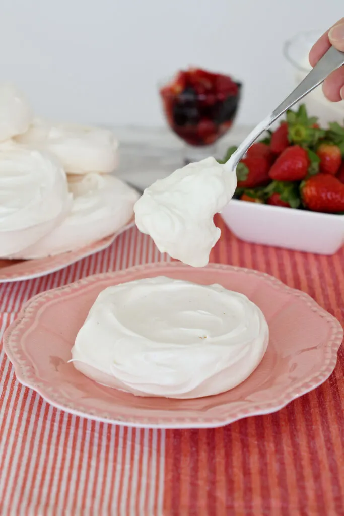 an individual pavlova on a pink plate and a spoon dropping whipped cream