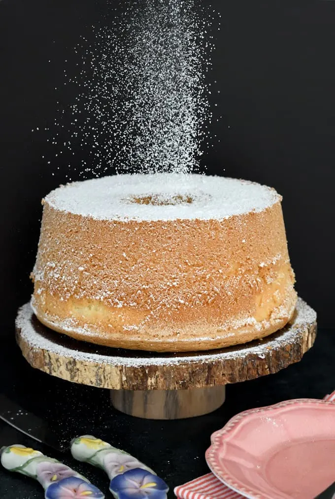 A vanilla chiffon cake on a cake stand being sprinkled with sugar