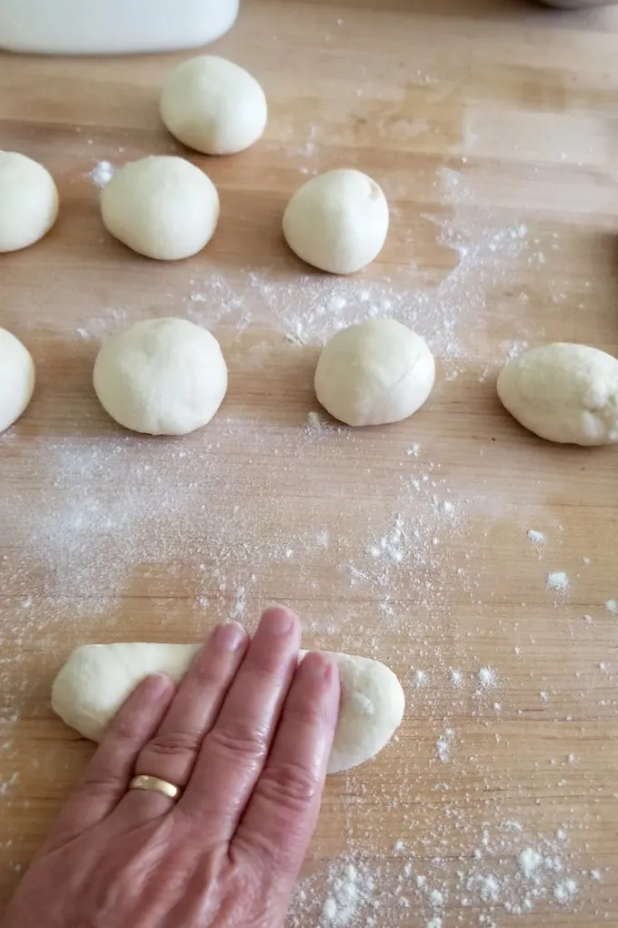 balls of hot dog bun dough on a wood surface. A hand is rolling a piece of dough into a cigar shape