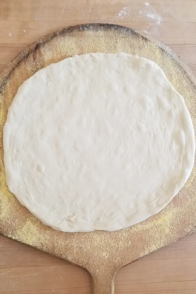an unbaked pizza crust on a wooden peel dusted with cornmeal