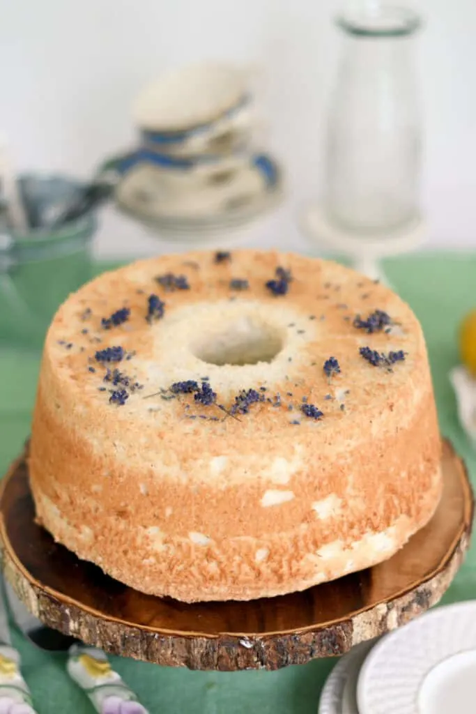 a lemon lavender angel food cake on a wooden cake stand. Lavender blossoms scattered on top of the cake