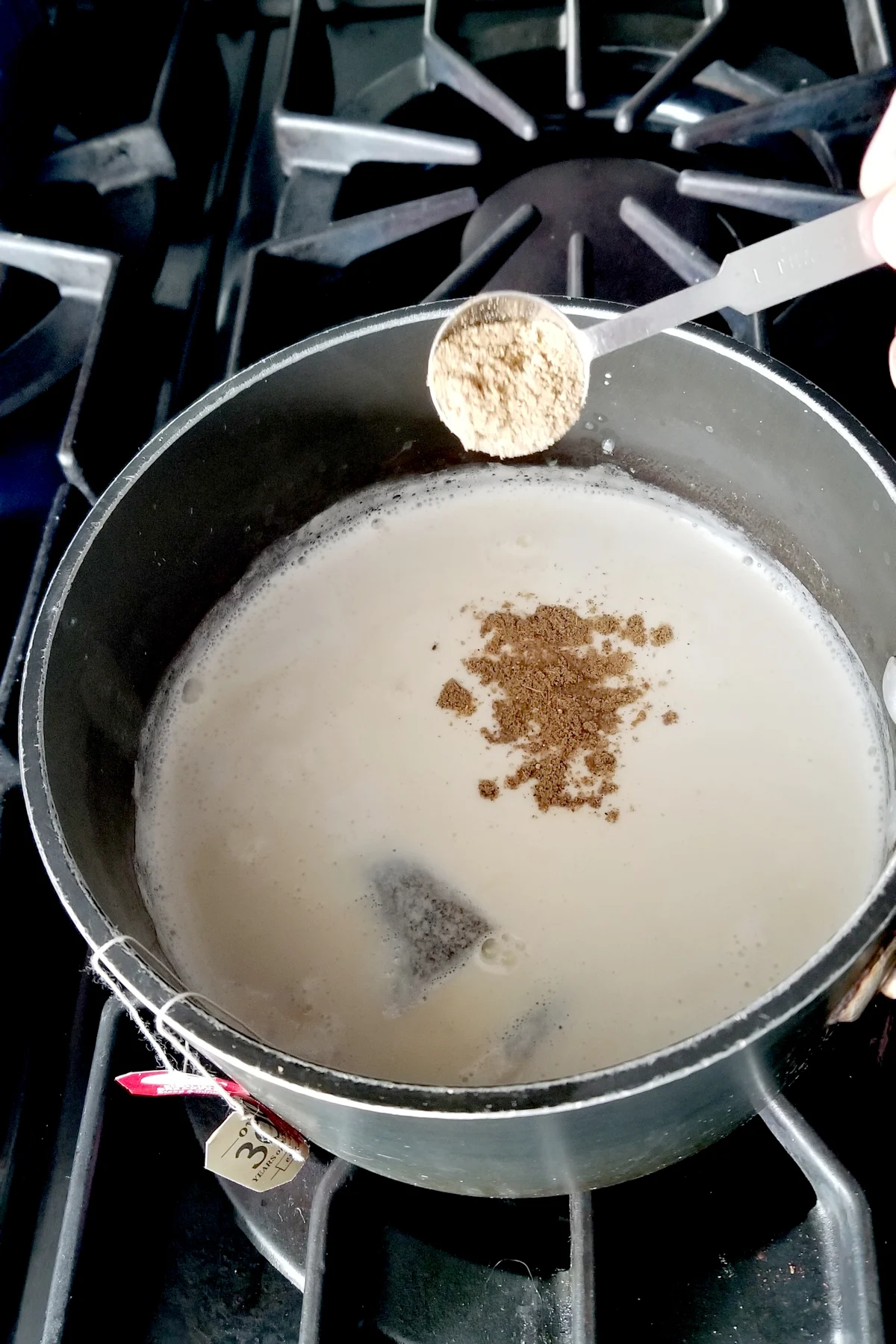 a pot of milk with tea bags steeping and a spoonful of chai spice being added to the mix.