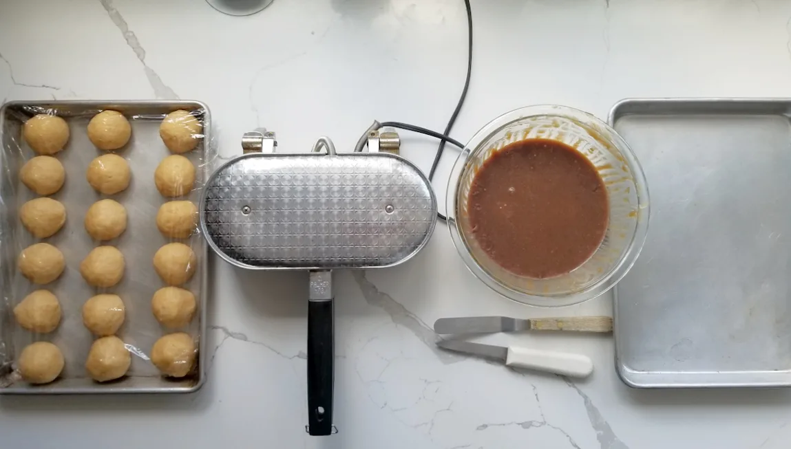a tray of cookie dough balls, a waffle iron, a bowl of caramel an empty sheet pan are set up on a countertop.