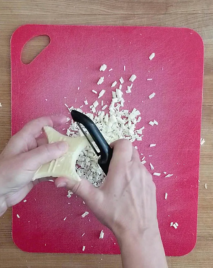 a red cutting board and hand peeling white chocolate shavings with a vegetable peeler