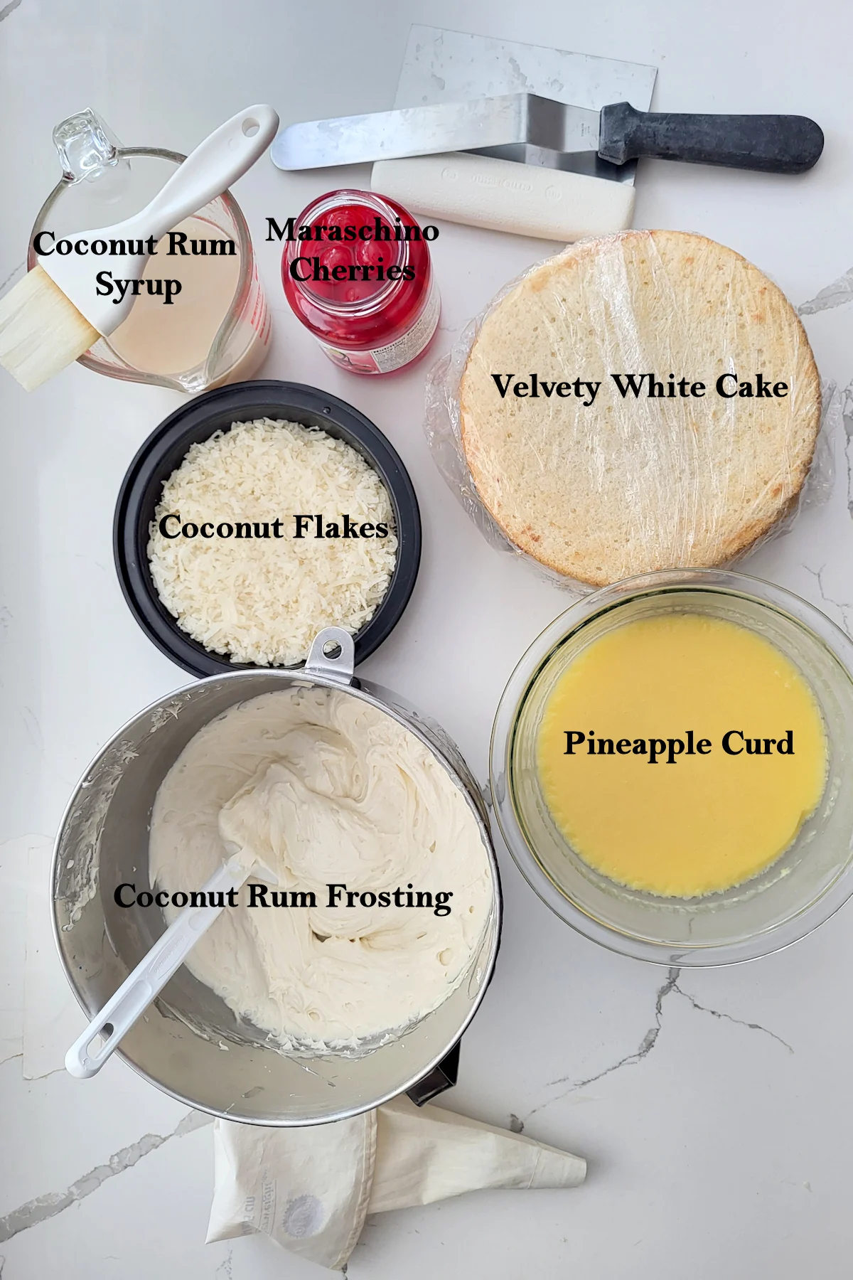 Cake layers, a bowl of coconut, a bowl of pineapple curd, a bowl of frosting, a jar of cherries, a piping bag, spatula, pastry brush and a scraper.