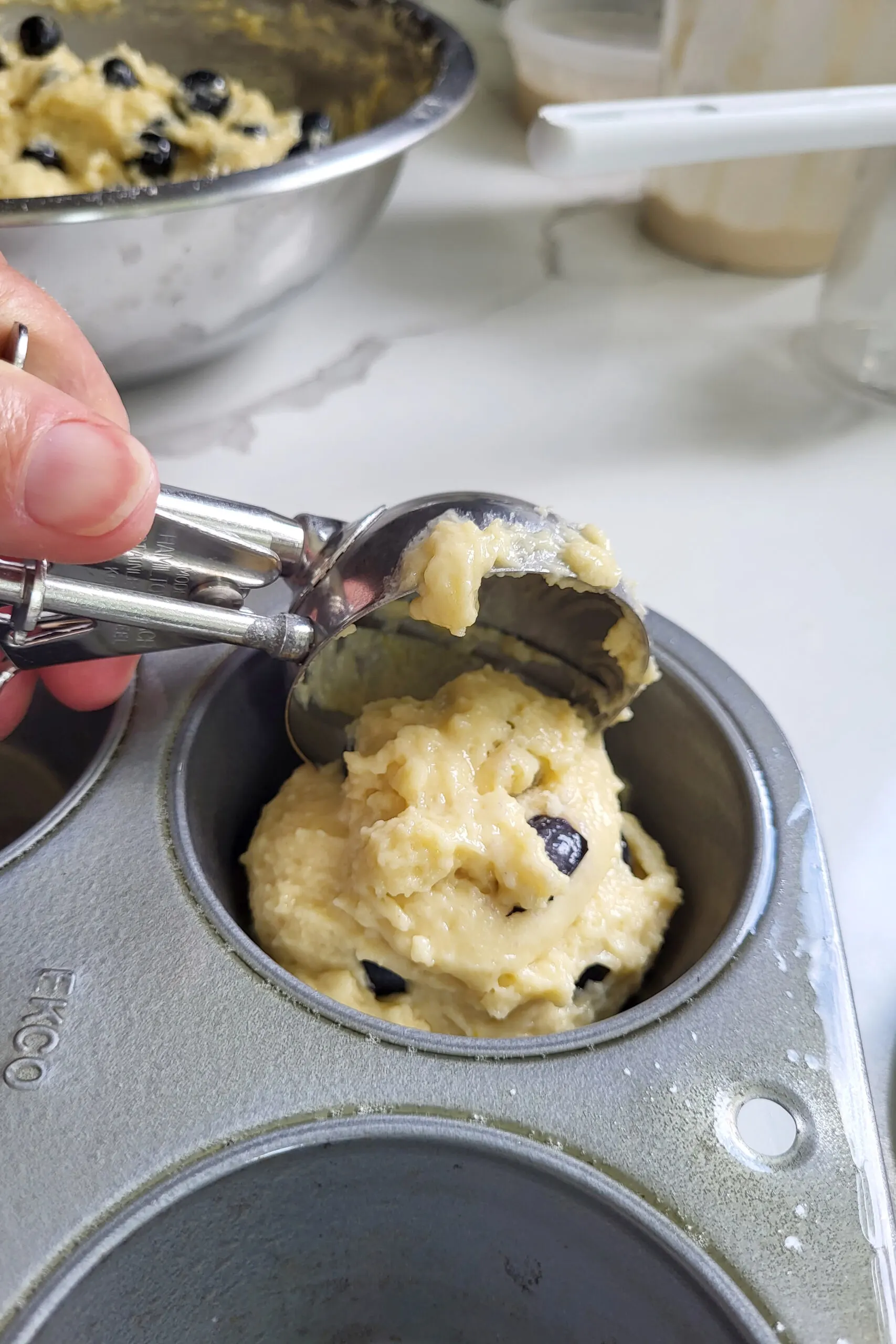 a scoop of blueberry muffin batter going into a greased muffin pan.