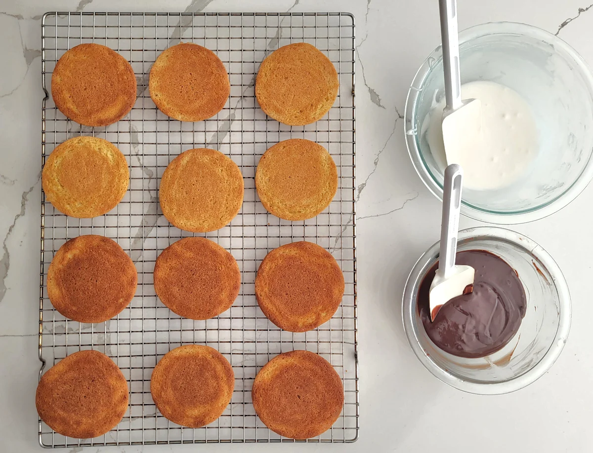 a cooling rack with baked cookies. A bowl of vanilla icing and a bowl of chocolate icing.