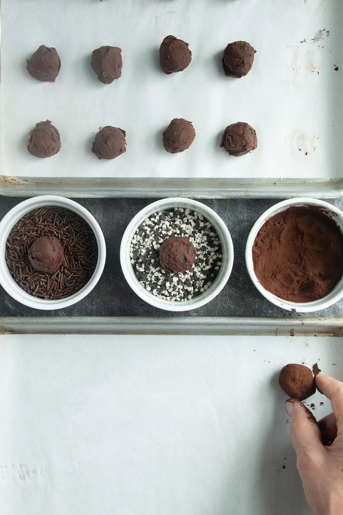 a tray of truffles, three bowls of sprinkles and a hand rolling a truffle in cocoa.