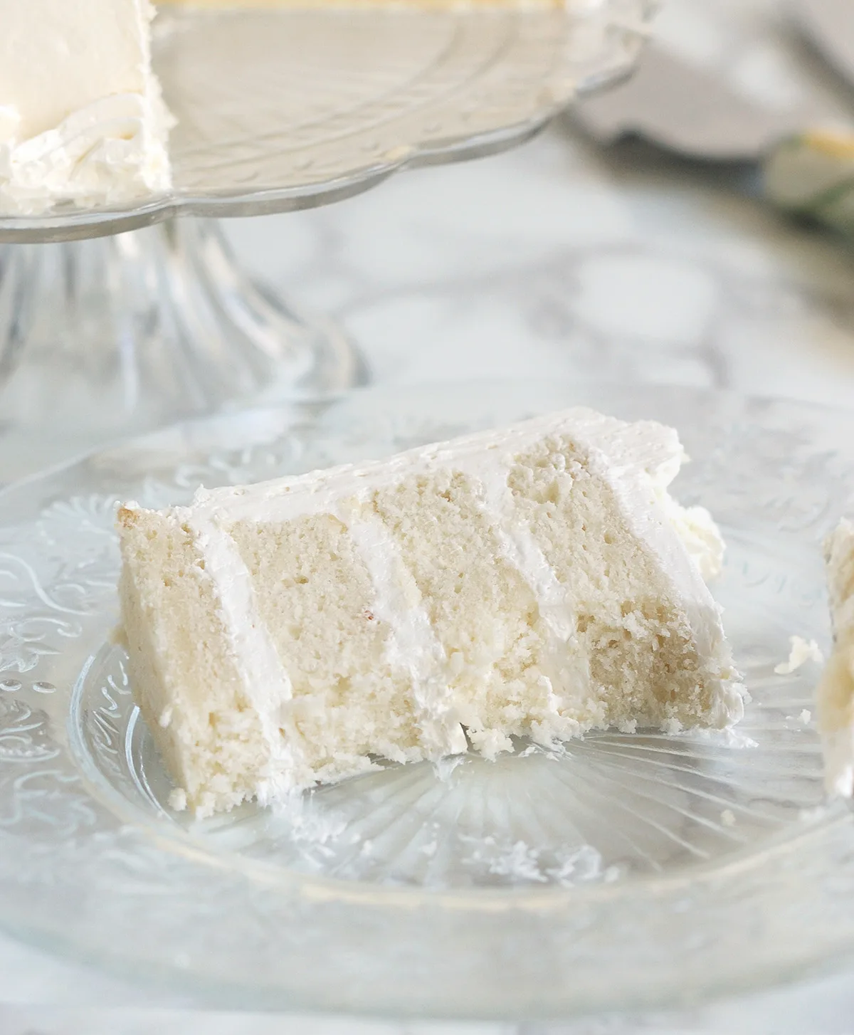 A half-eaten slice of four layer velvety soft white cake on a glass plate
