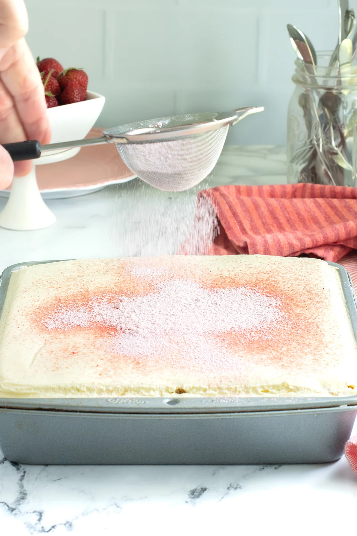A sifter sprinkling strawberry sugar over a tiramisu in a pan.