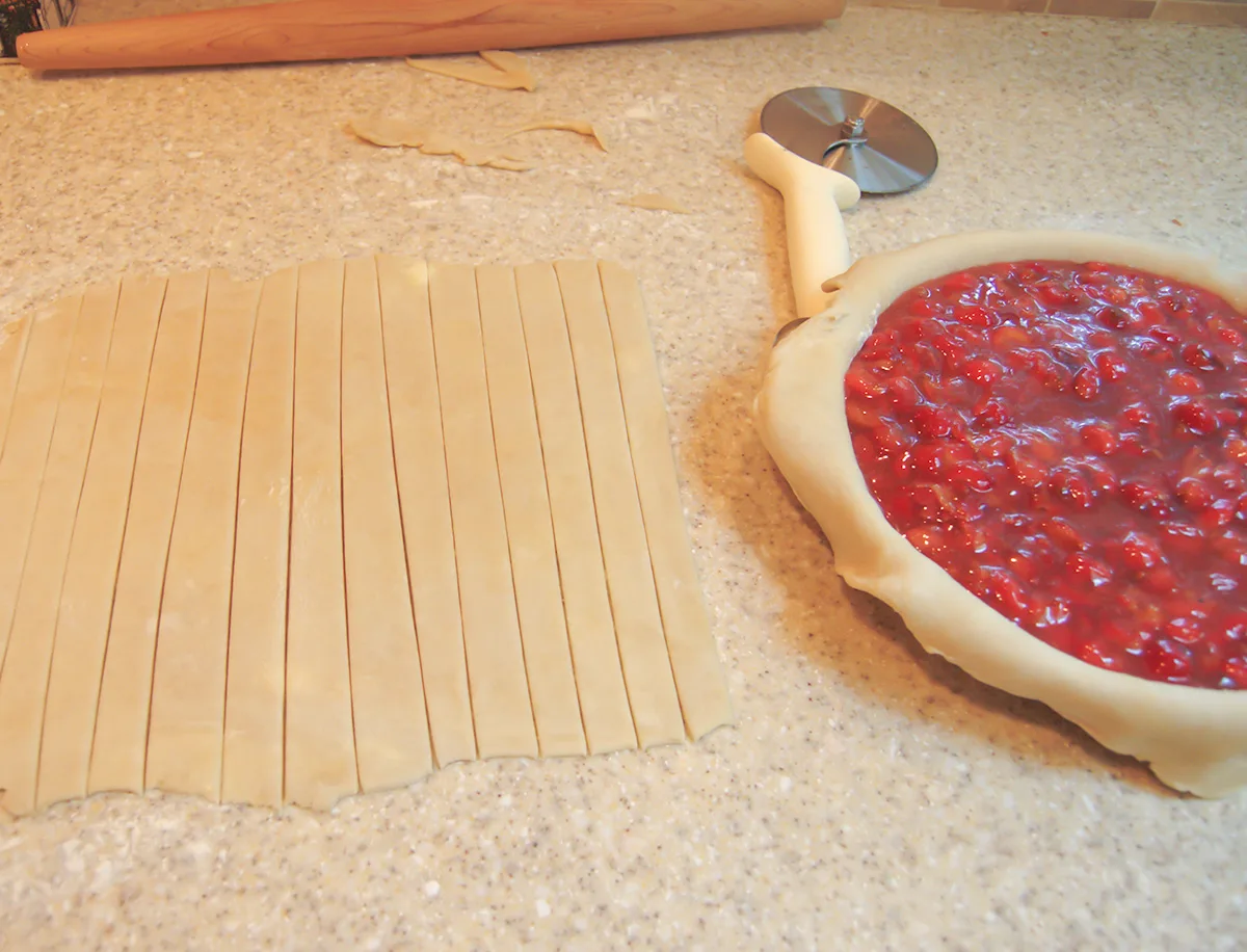 A cherry pie without a top. Strips of pie dough on the counter.