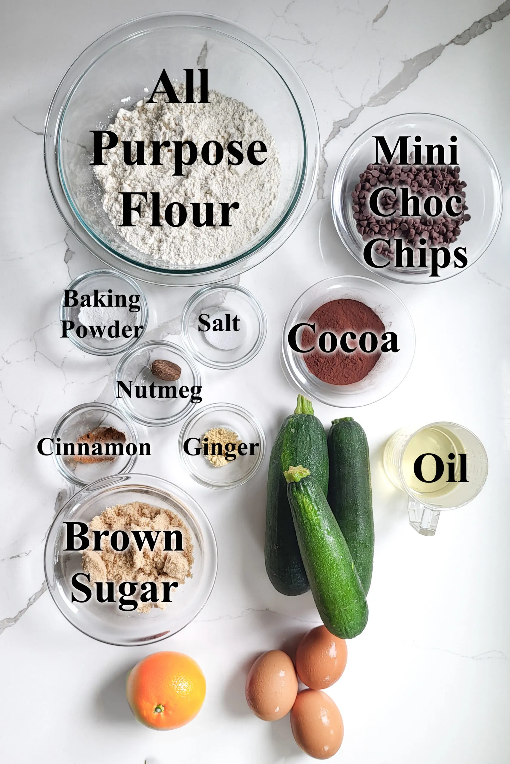 ingredients for chocolate orange zucchini bread in glass bowls on a white surface.