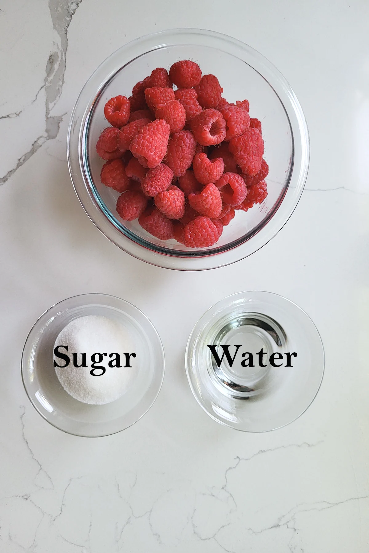 ingredients for raspberry compote in glass bowls on a white surface.