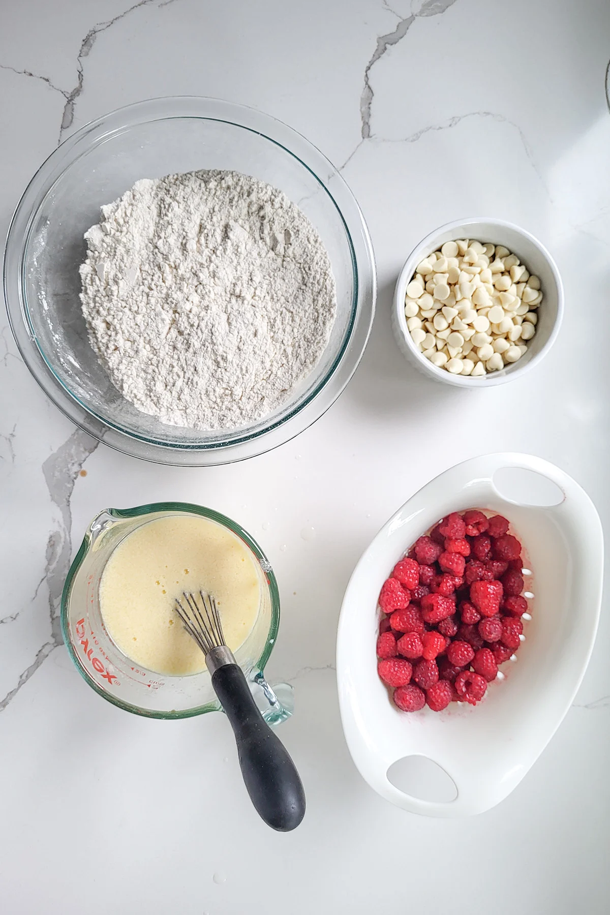 A bowl of flour, a cup with milk and eggs, a bowl of white chocolate chips and a basket of raspberries.