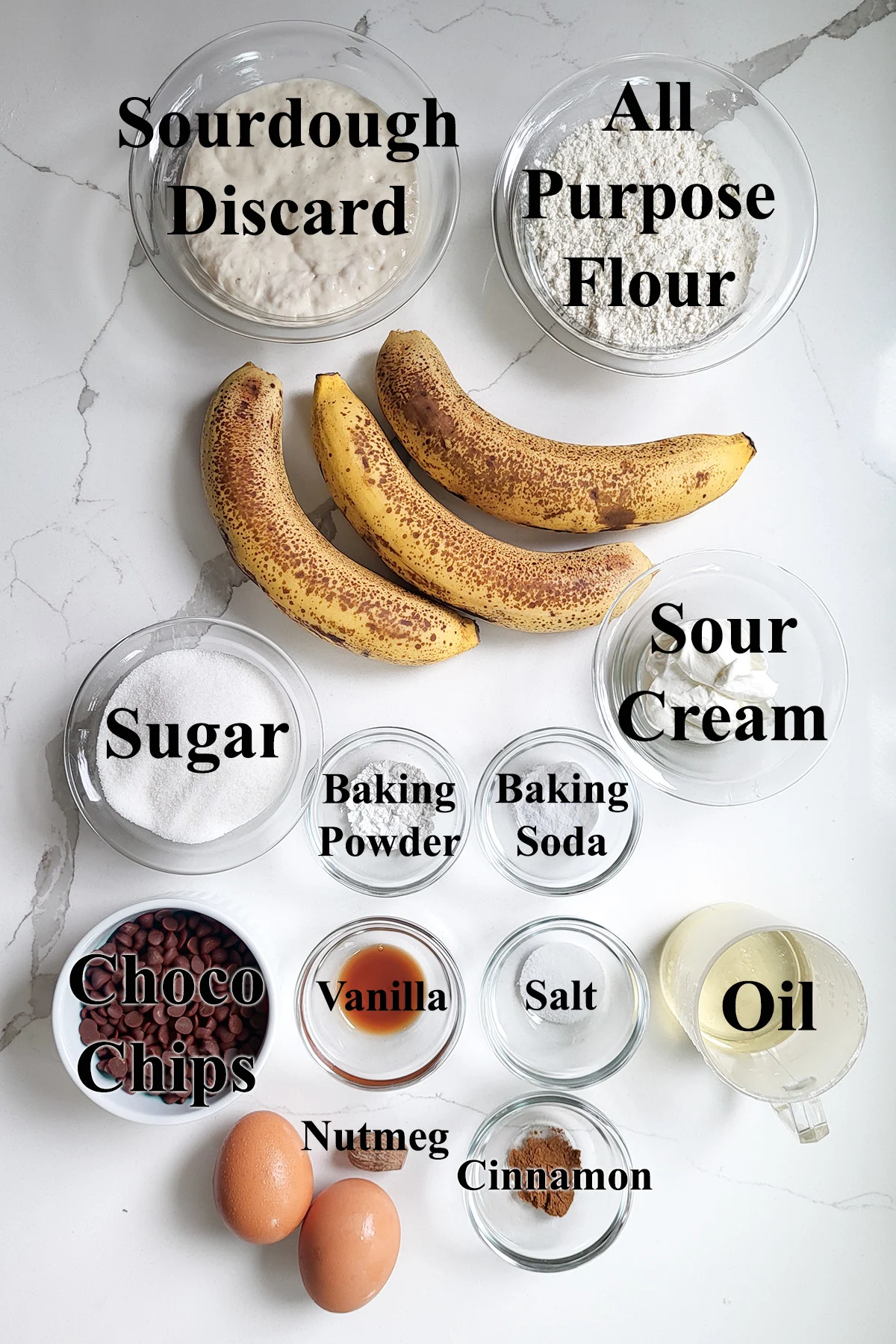 ingredients for sourdough banana bread in glass bowls on a white surface.