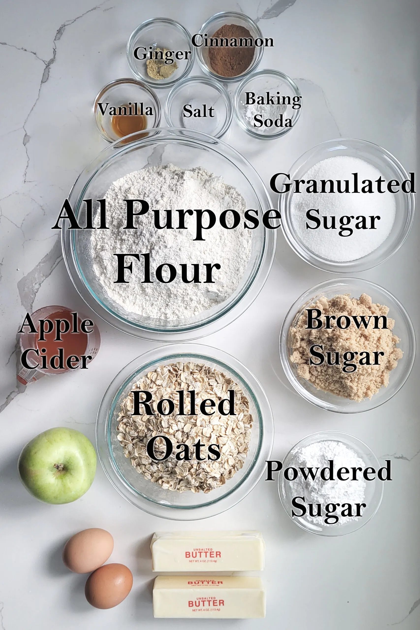 ingredients for apple oatmeal cookies in glass bowls on a white surface.