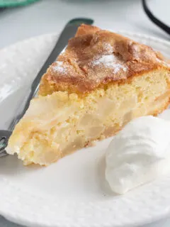 A slice of pear cake, a dollop of whipped cream and a fork on a white plate.