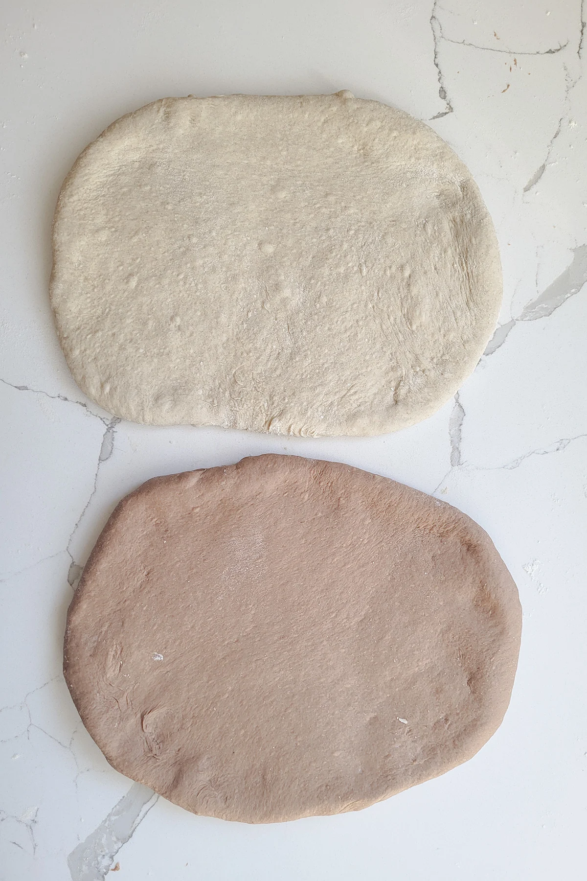 an oval or light bread dough and an oval of dark bread dough on a marble surface.