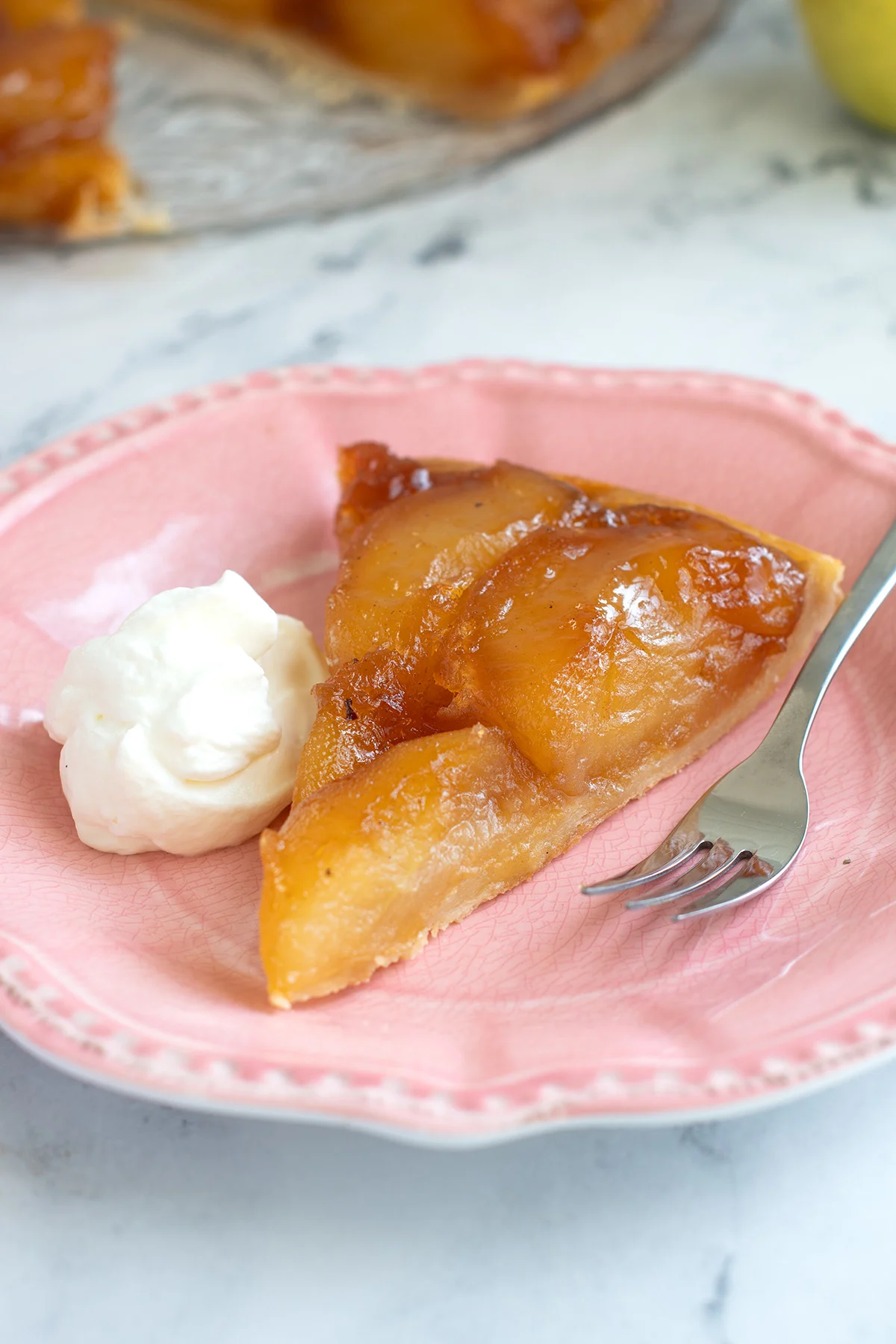a slice of tarte tatin on a pink plate with a fork and a dollop of cream.