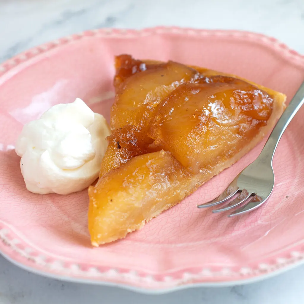 a slice of tarte tatin on a pink plate with a fork and dollop of whipped cream.