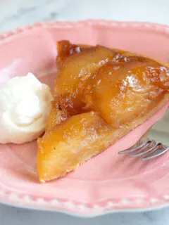 a slice of tarte tatin on a pink plate with a fork and dollop of whipped cream.