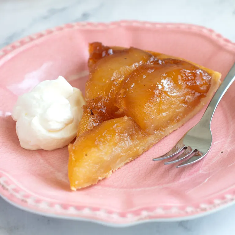 a slice of tarte tatin on a pink plate with a fork and dollop of whipped cream.