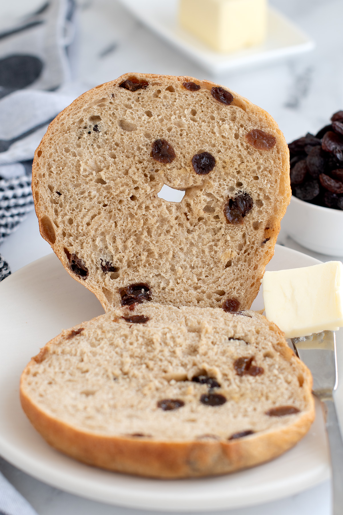 a sliced cinnamon raisin bagel on a white plate with a pat of butter. 