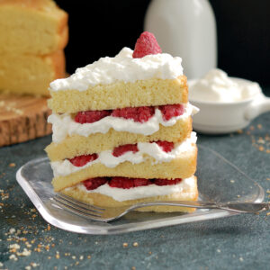 a slice of genoise cake with raspberries and cream on a glass plate.