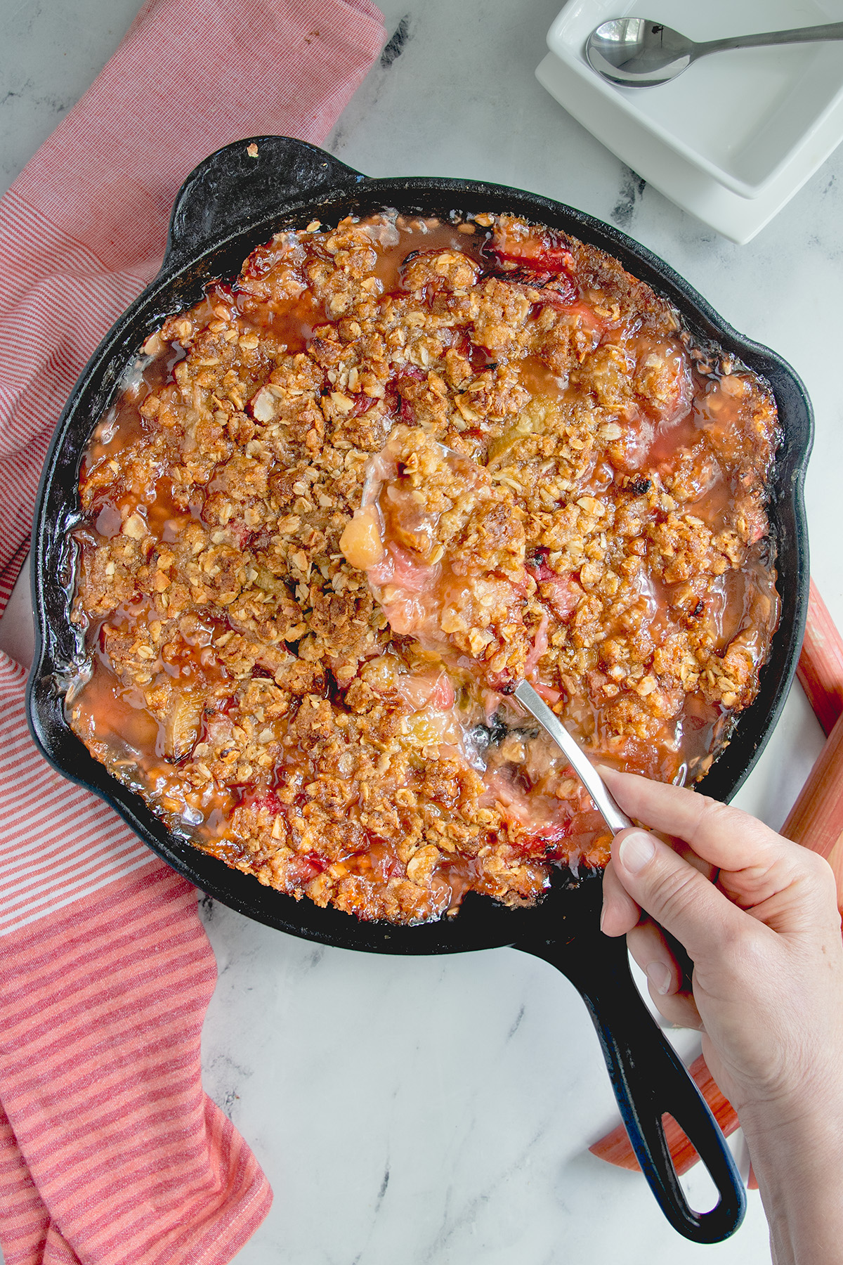 a cast iron pan filled with rhubarb crisp and a hand with a spoon.